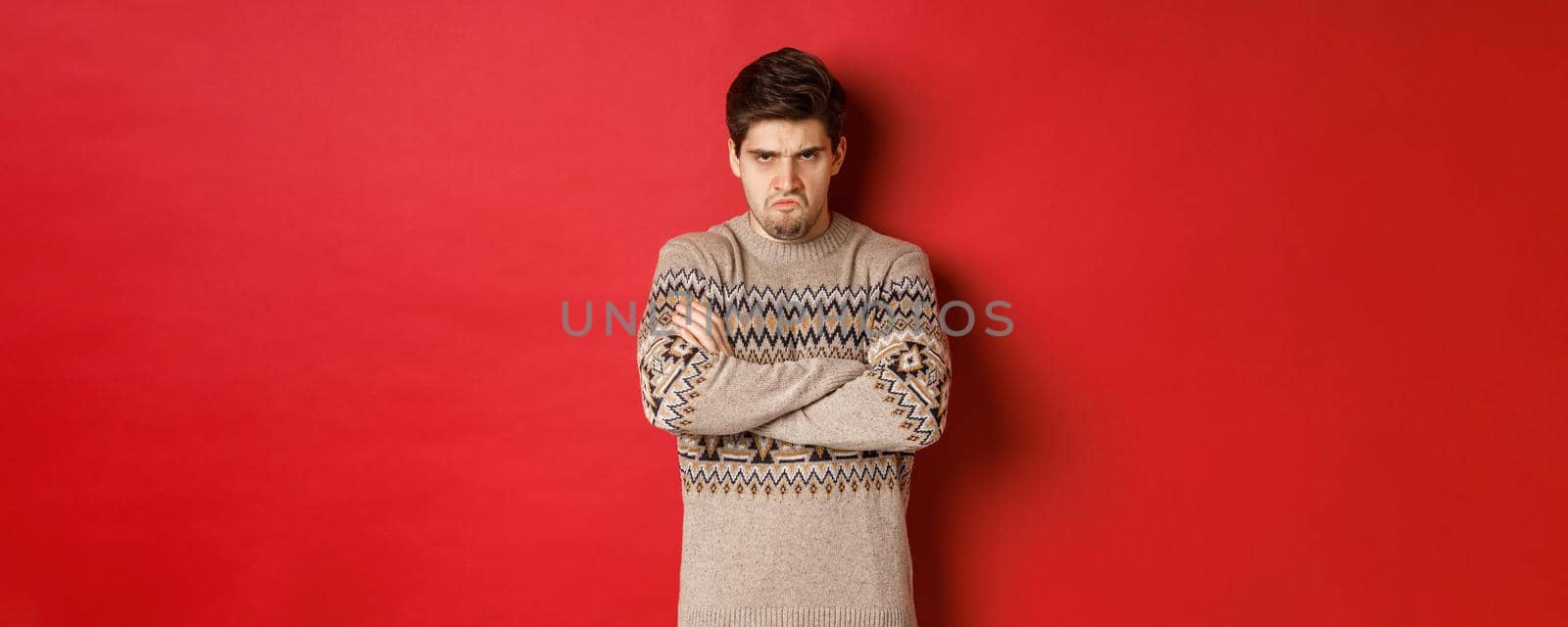 Image of angry and offedned guy in christmas sweater, feeling mad, cross arms on chest and sulking, standing over red background by Benzoix
