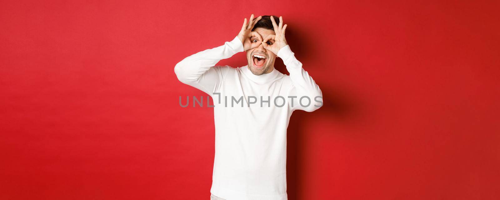 Portrait of handsome guy in white sweater, making funny mask with fingers, looking happy and smiling, standing over red background.