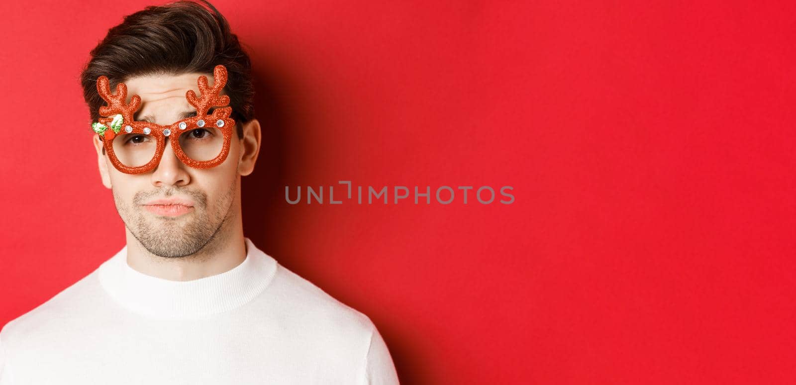 Concept of winter holidays, christmas and celebration. Close-up of skeptical handsome guy, wearing party glasses, smirk and looking unamused, standing over red background.