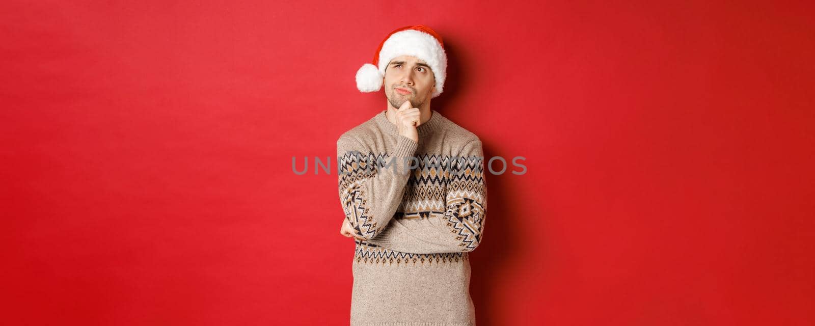 Image of attractive man in christmas sweater and santa hat, thinking about new year gifts, looking at upper left corner thoughtful, standing over red background.