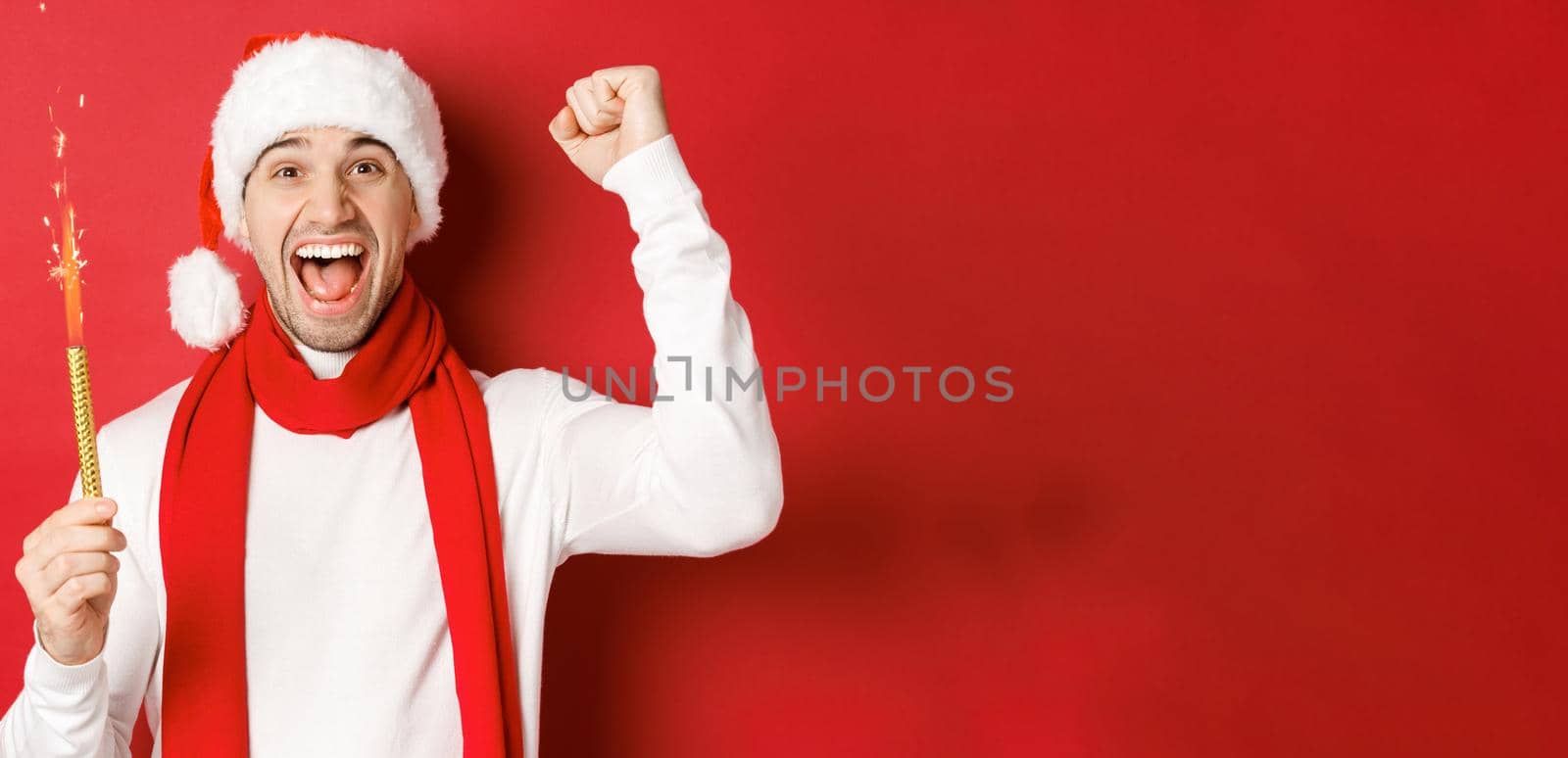 Concept of christmas, winter holidays and celebration. Handsome man celebrating new year and having fun, holding sparkler and smiling, wearing santa hat, standing over red background by Benzoix