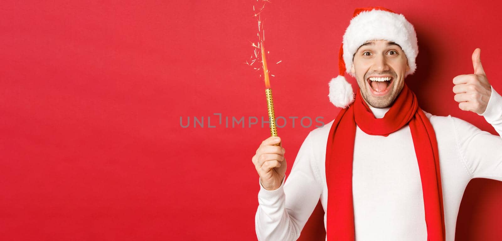 Concept of christmas, winter holidays and celebration. Handsome man celebrating new year and having fun, holding sparkler and showing thumb-up, standing over red background.