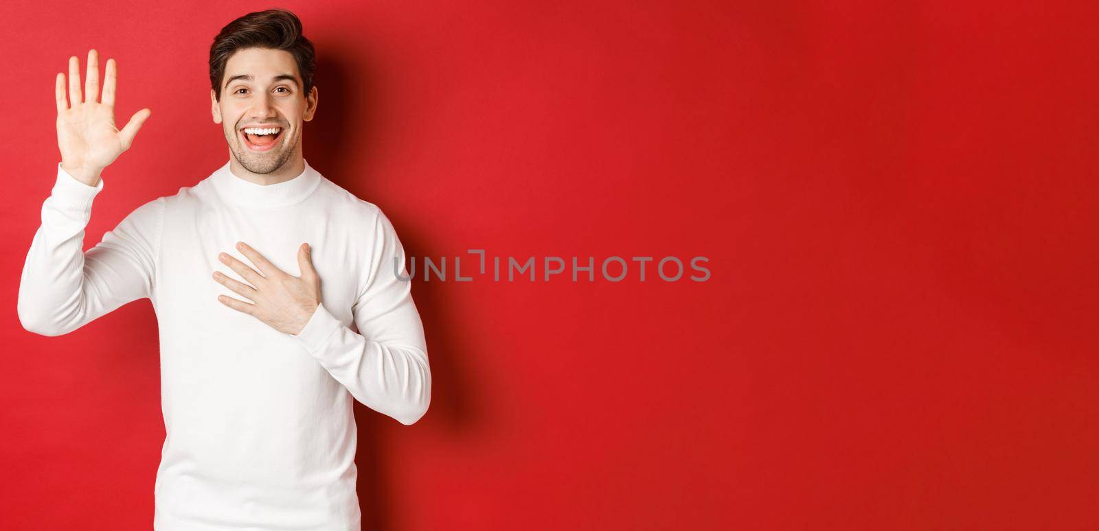 Portrait of honest smiling man in white sweater, making a promise, swearing to tell truth, standing against red background.