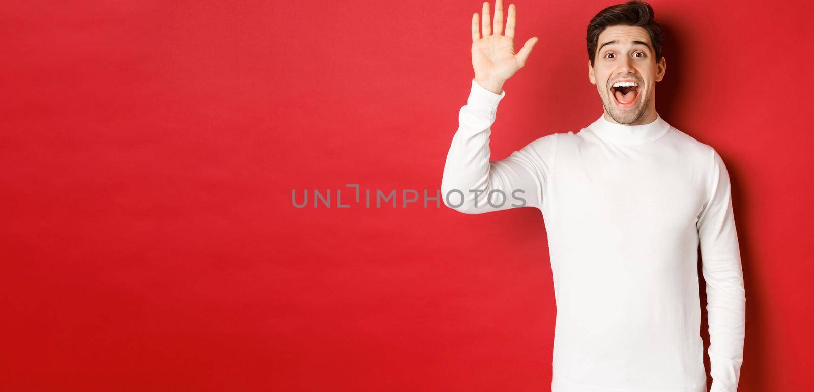Image of happy and friendly young man saying hello, waving hand to greet someone, standing over red background.