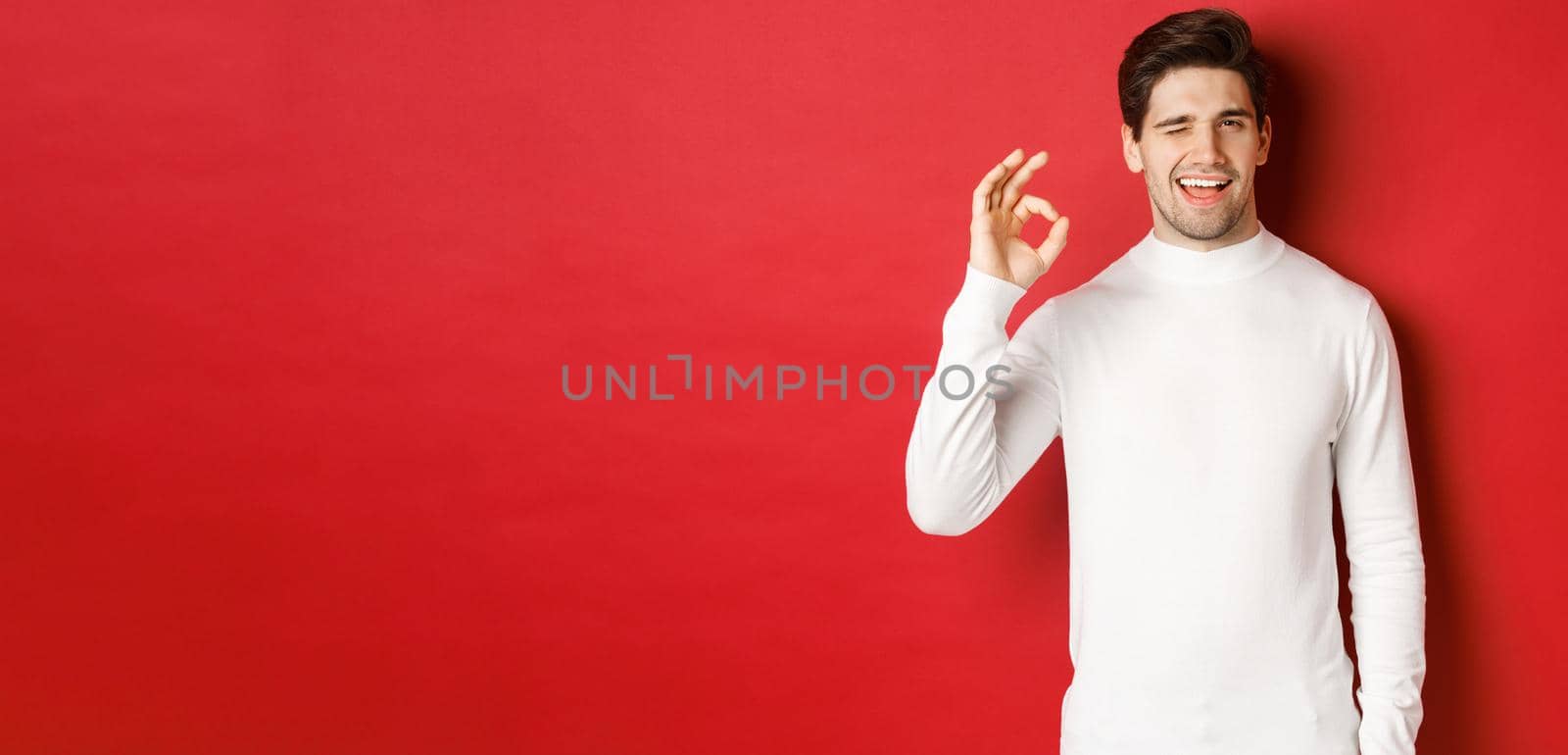 Portrait of charismatic beaded guy in white sweater, assure you, winking and showing okay sign to guarantee good quality, praise excellent choice, standing over red background by Benzoix