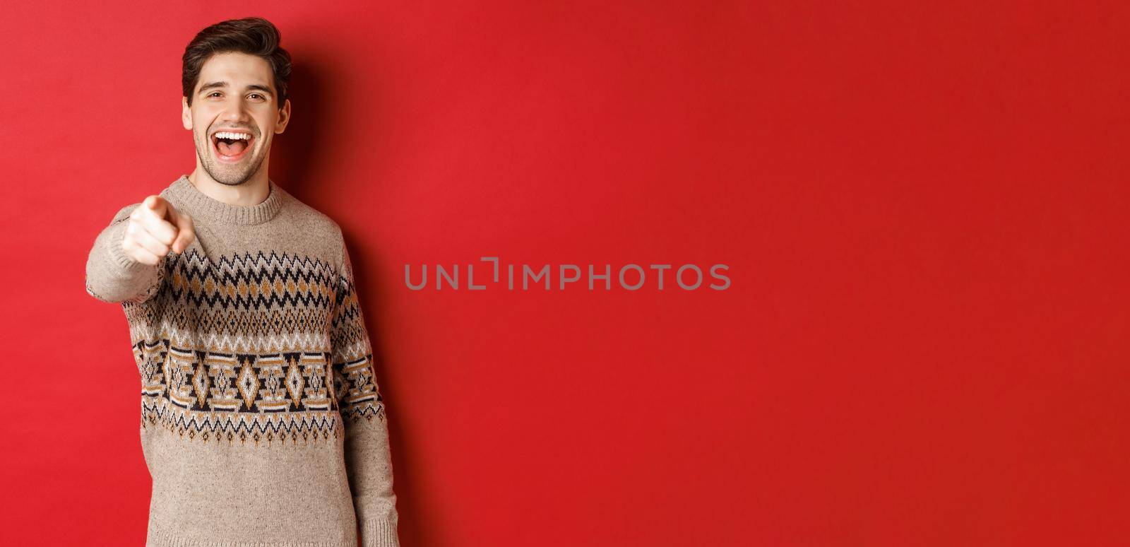 Concept of christmas celebration, winter and lifestyle. Happy attractive man in xmas sweater, wishing happy holidays and pointing at camera, standing over red background.