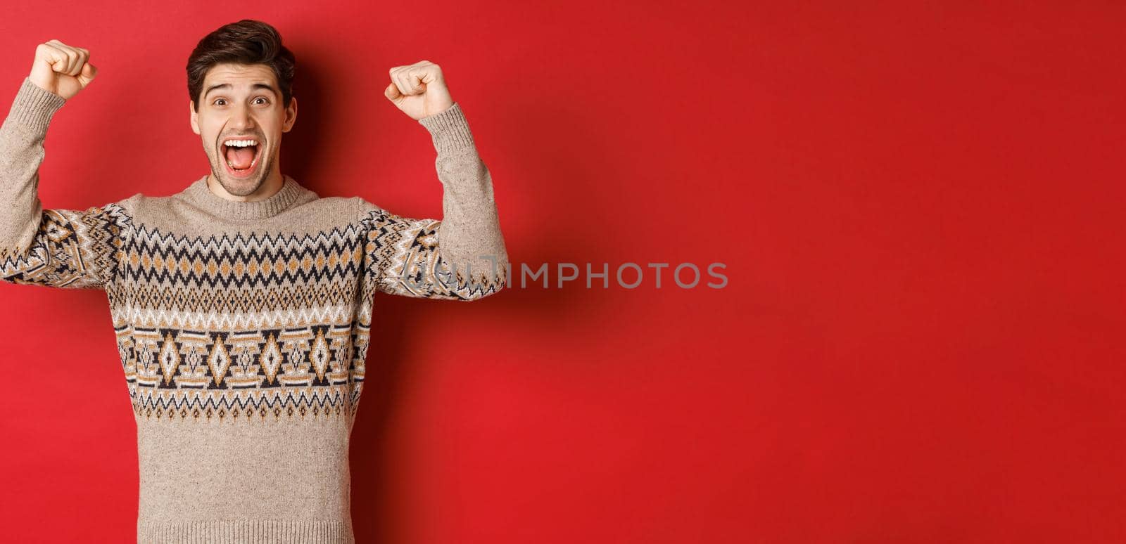 Image of happy handsome guy rejoicing on christmas eve, raising hands up and shouting for joy yes, achieve new year goal, standing over red background in sweater.