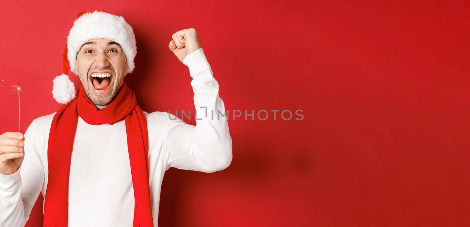 Concept of christmas, winter holidays and celebration. Portrait of excited handsome man, raising hand up and holding sparkler, wishing happy new year, standing over red background by Benzoix