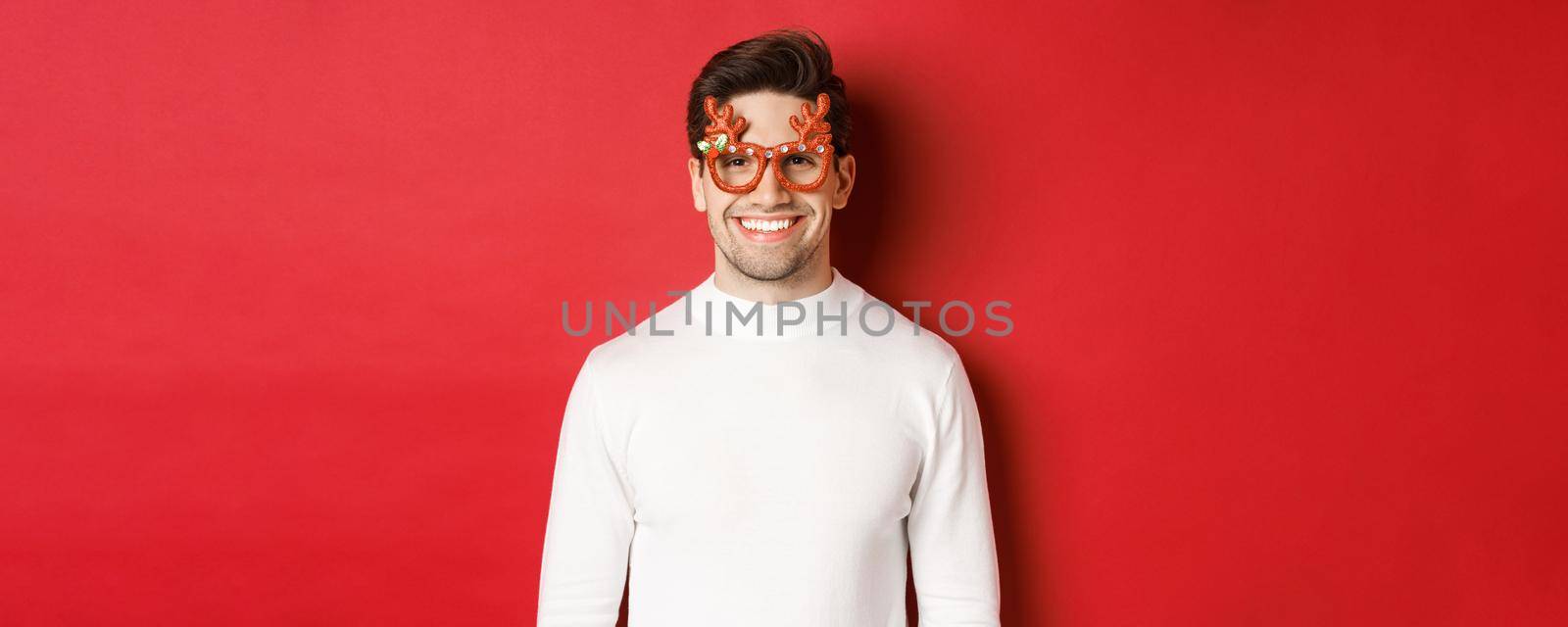 Close-up of attractive man in white sweater and party glasses, celebrating christmas, smiling amused, standing over red background by Benzoix