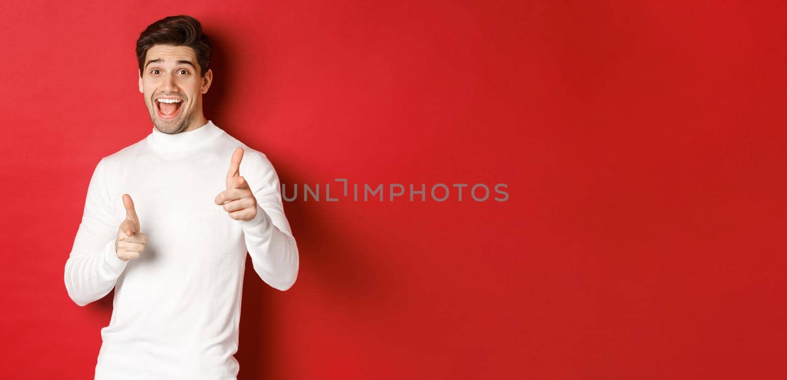 Concept of winter holidays, christmas and lifestyle. Cheerful handsome guy in white sweater congratulating you, pointing fingers at camera and wishing happy new year, standing over red background by Benzoix