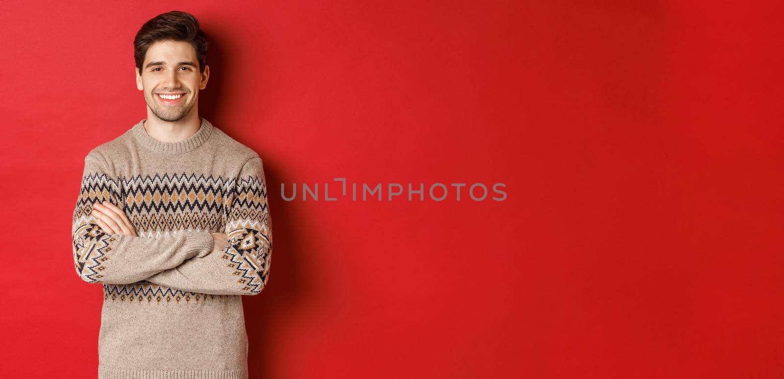 Image of handsome happy guy in christmas sweater, smiling and looking at camera, celebrating xmas holidays, standing over red background.