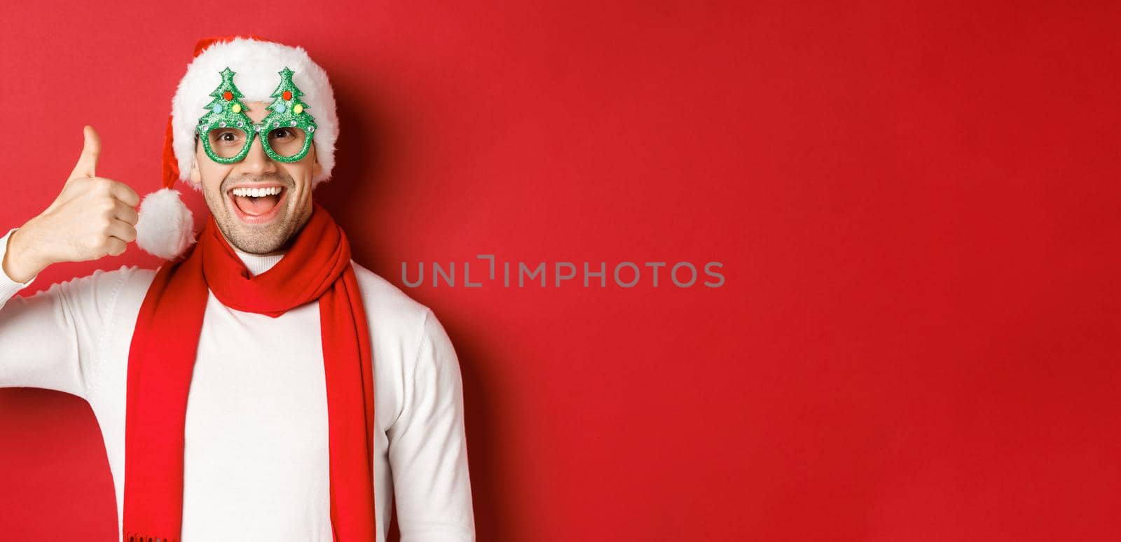 Concept of christmas, winter holidays and celebration. Cheerful guy in santa hat and party glasses, showing thumb-up and smiling pleased, standing over red background by Benzoix