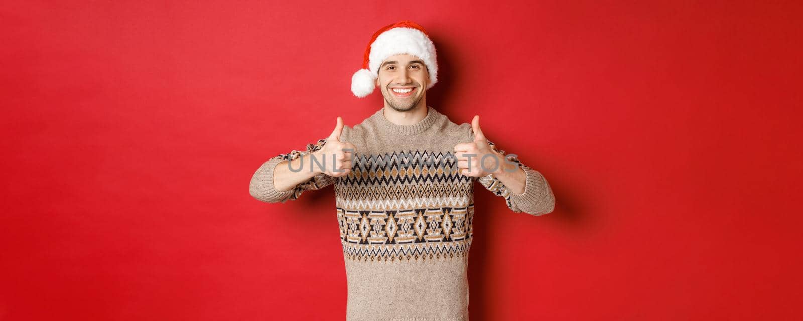 Image of cheerful attractive man in winter sweater and santa hat, showing thumbs-up, celebrating christmas and looking happy, like something, standing over red background.