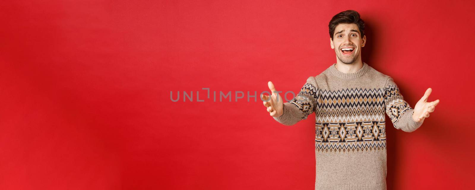 Image of happy and flattered handsome guy in christmas sweater, reaching hands to welcome guests on new year party, inviting to celebration, standing over red background.