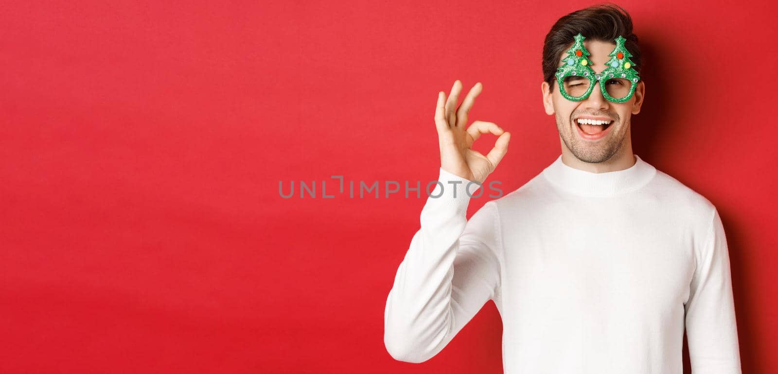 Portrait of handsome and cheeky guy, wearing party glasses and white sweater, showing okay sign and winking, wishing happy new year, standing over red background.