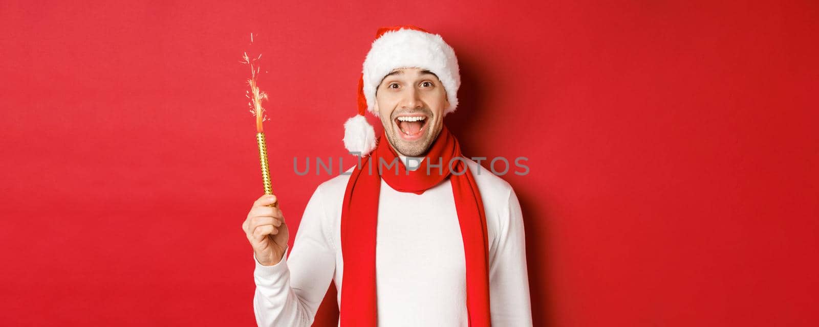 Concept of christmas, winter holidays and celebration. Handsome man celebrating new year and having fun, holding sparkler and smiling, wearing santa hat, standing over red background.