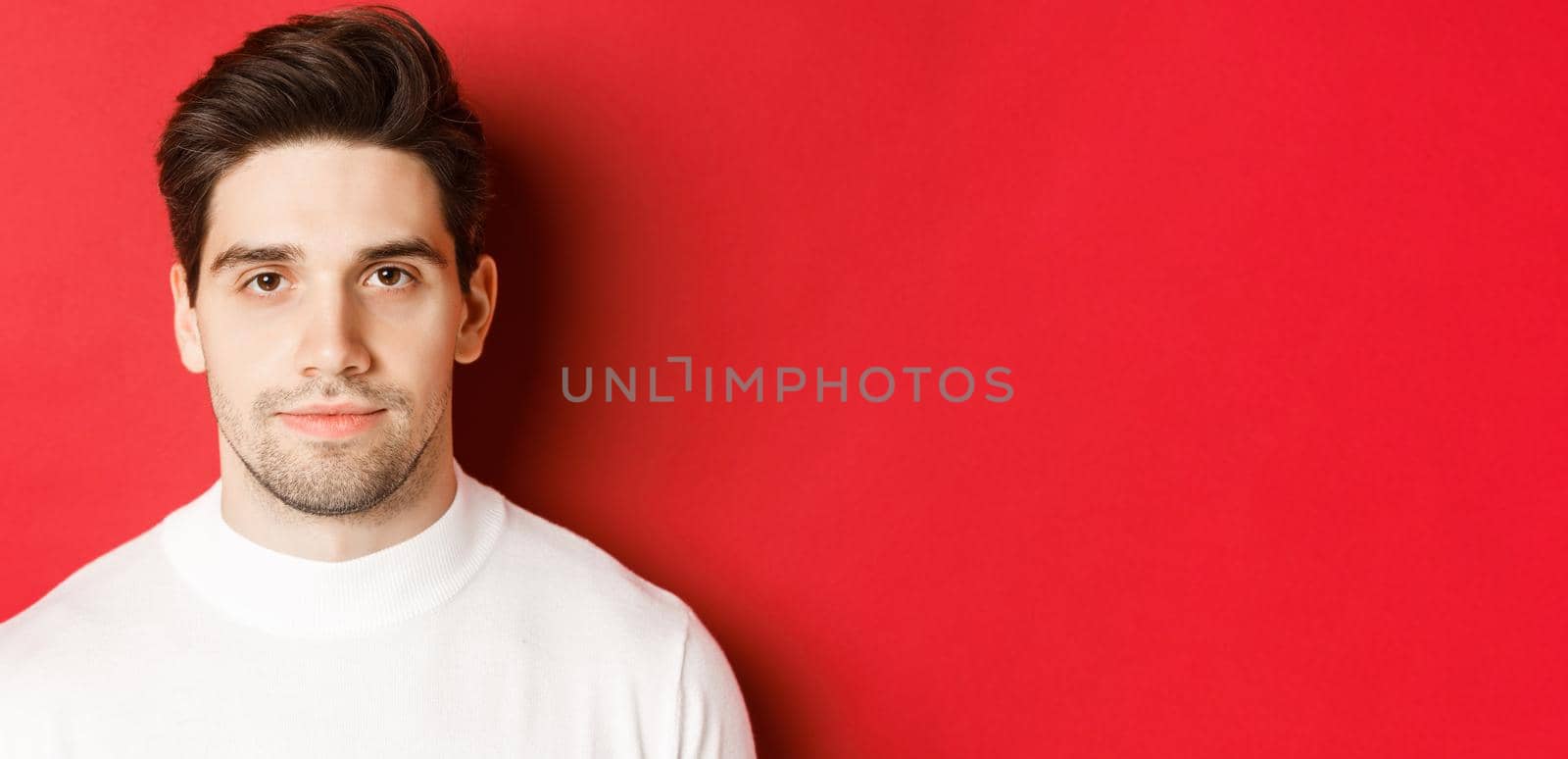 Close-up of handsome brunette guy in white sweater, smiling at camera, standing over red background.