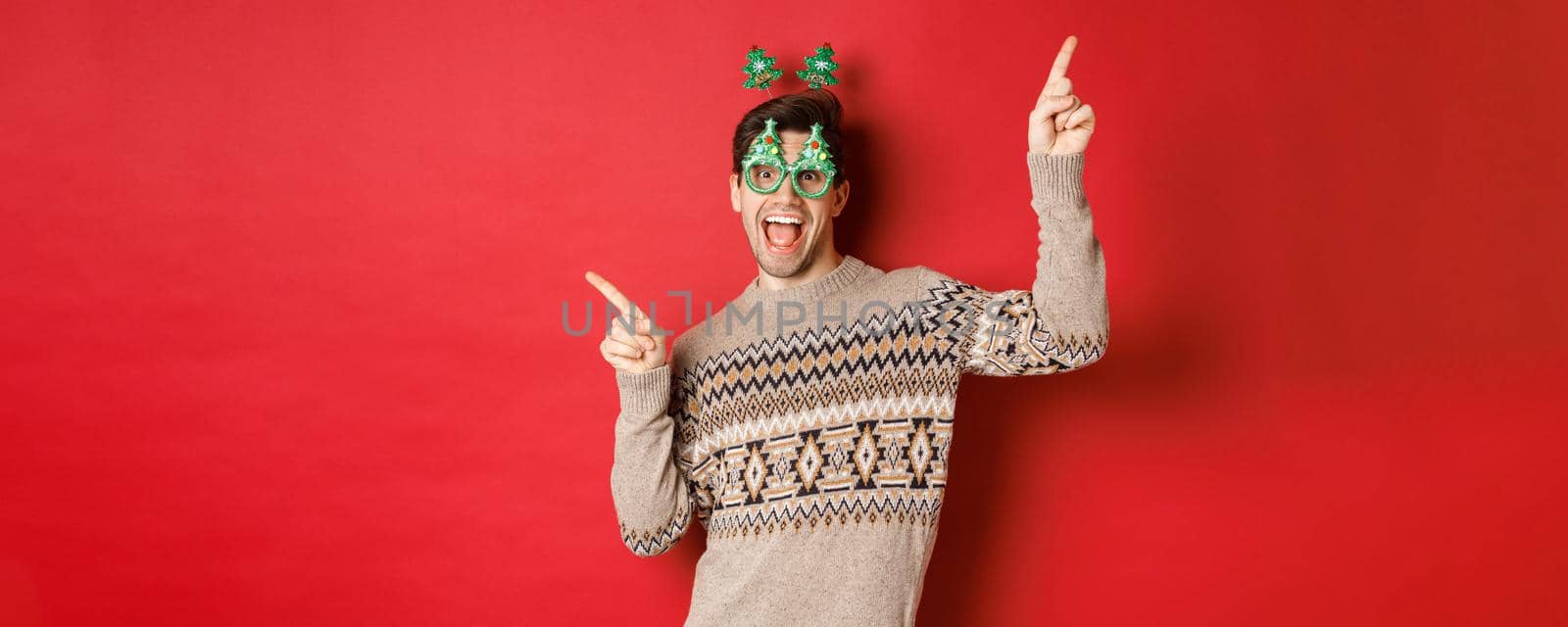 Portrait of joyful handsome guy in party glasses and christmas sweater, dancing and pointing fingers sideways, enjoying new year celebration, standing over red background by Benzoix
