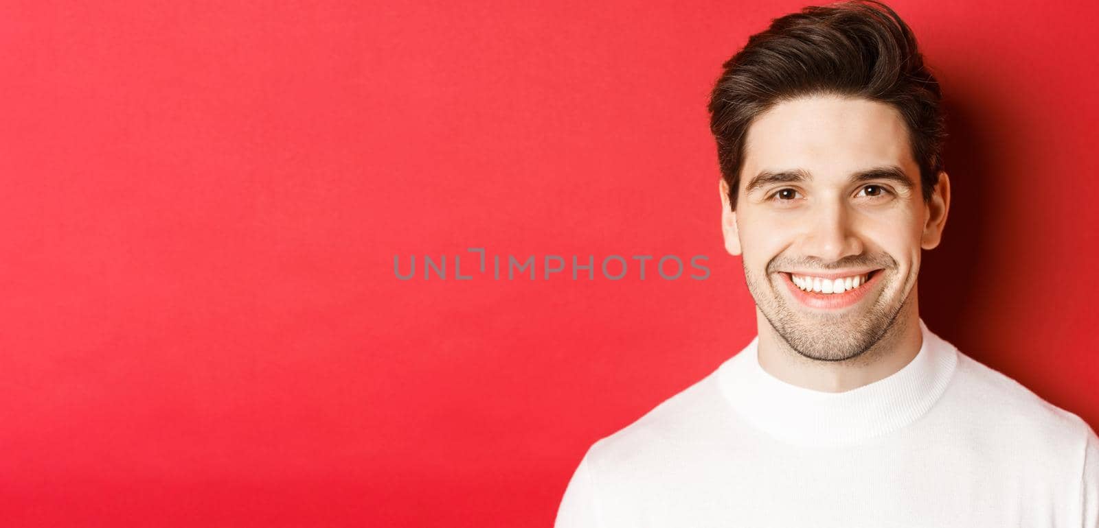 Close-up of handsome smiling brunette man, wearing white sweater, smiling happy and confident, standing against red background.