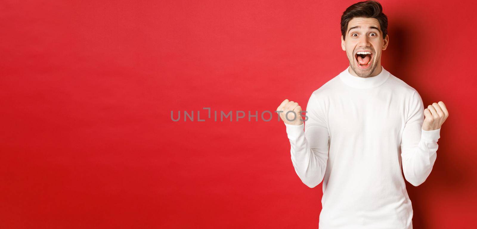 Image of happy good-looking man in white sweater, winning something, making fist pump and smiling amazed, celebrating victory, standing over red background.