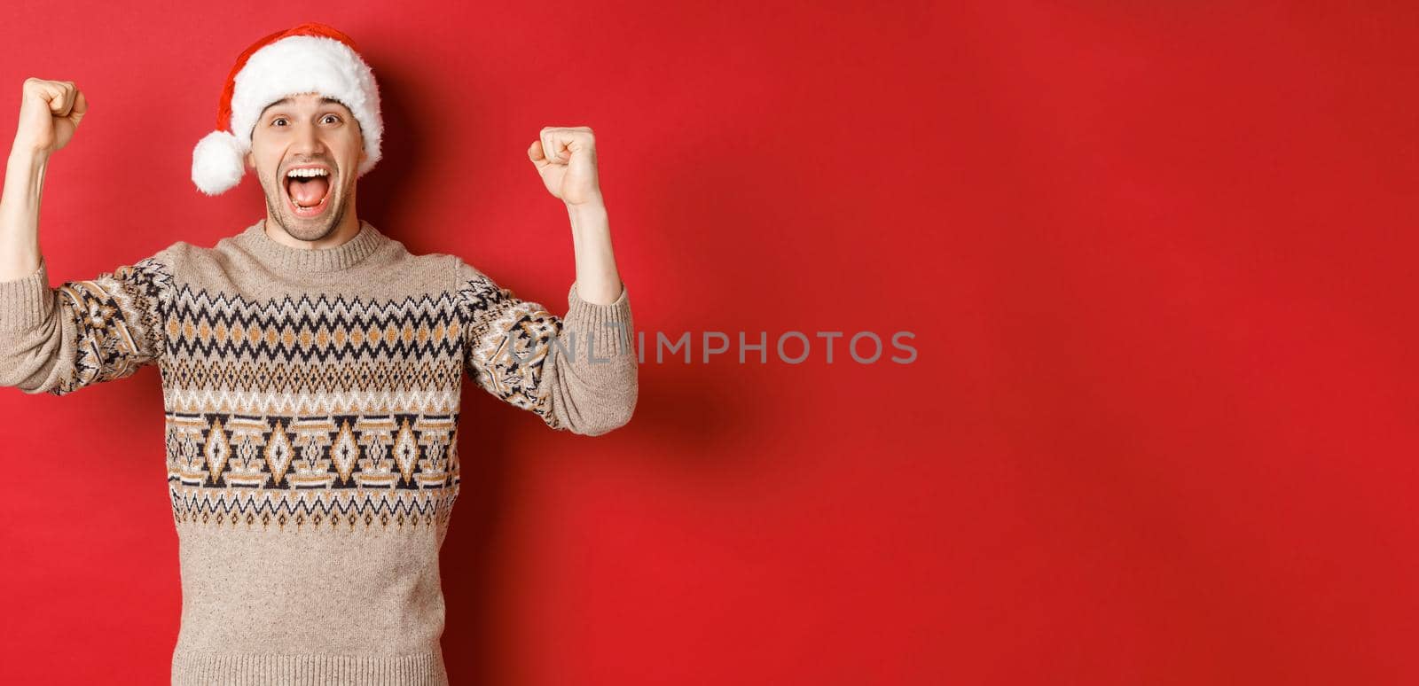 Image of cheerful handsome man in swearer and santa hat, celebrating new year, triumphing or winning something, raising hands up and shouting for joy, standing over red background by Benzoix