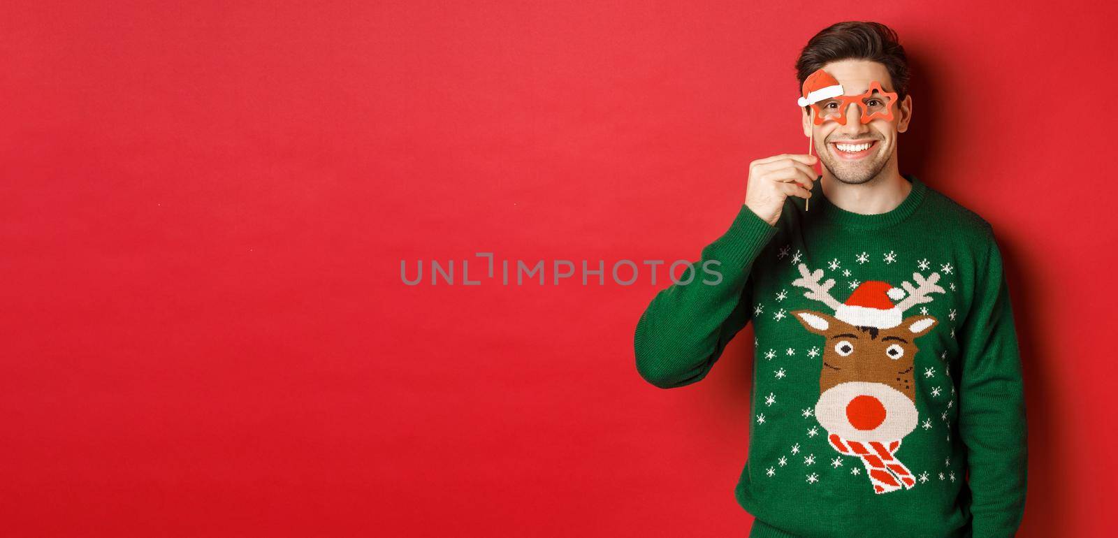 Portrait of handsome smiling man in christmas sweater and party glasses, celebrating new year and having fun, standing against red background.