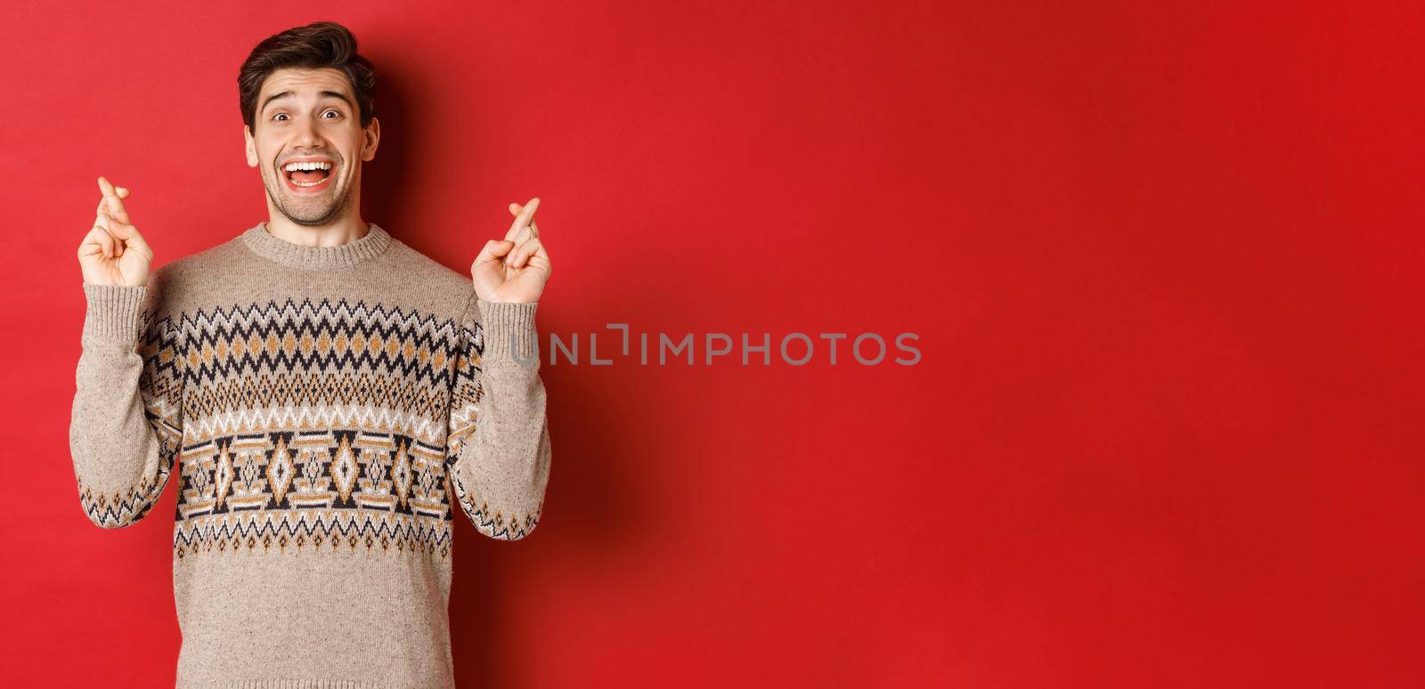 Image of excited and hopeful young man making wish, wearing winter sweater, crossing fingers for good luck and smiling, anticipating christmas eve, standing over red background.