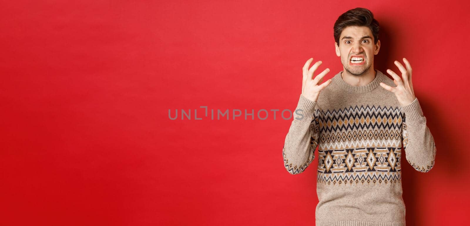 Image of angry and frustrated man in winter sweater, shouting and looking with hatred, standing upset over red background.