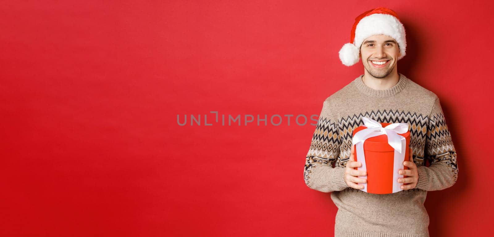 Portrait of handsome man holding a present, wishing happy holiday, standing in santa hat and christmas sweater against red background.