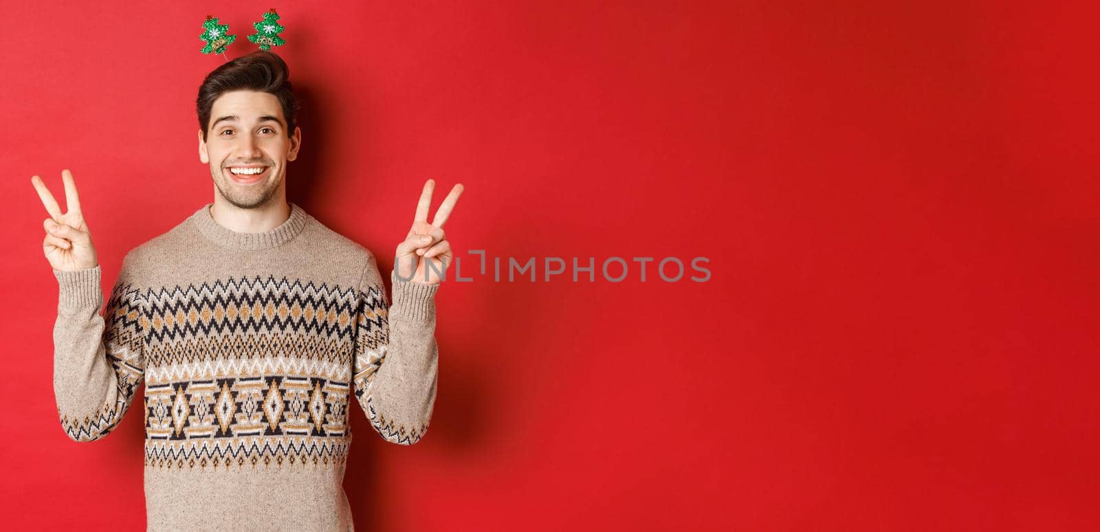 Concept of winter holidays, christmas and celebration. Image of handsome and silly guy dressed for new year party, showing peace signs and smiling, standing against red background.