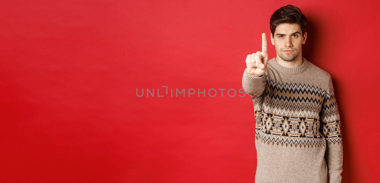 Image of serious handsome man extending one finger to stop you, telling no, give warning, standing over red background in winter sweater.