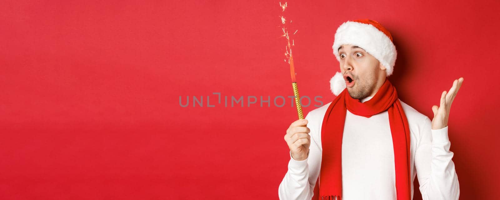 Concept of christmas, winter holidays and celebration. Close-up of amazed handsome man in santa hat and scarf, looking at sparkler with excitement, standing over red background by Benzoix