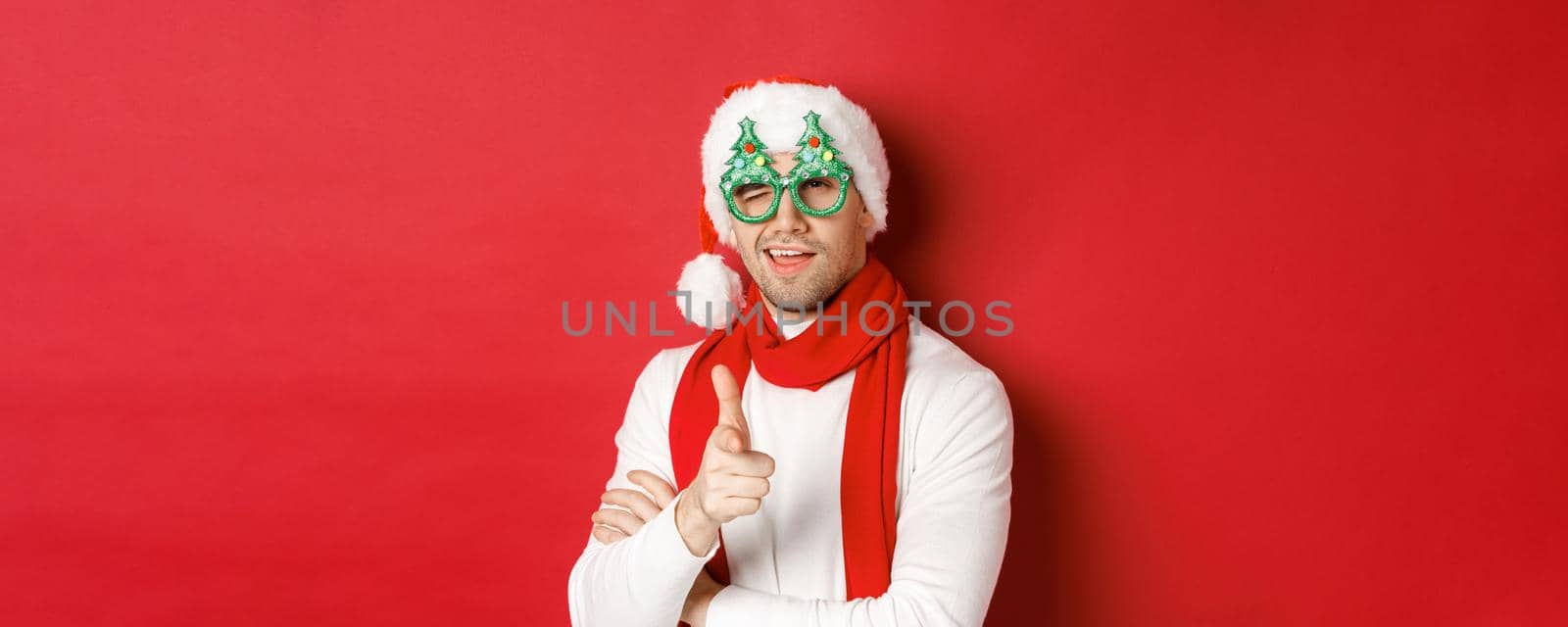 Concept of christmas, winter holidays and celebration. Close-up of cheeky young man in santa hat and party glasses, smiling and pointing finger gun at camera, standing over red background by Benzoix