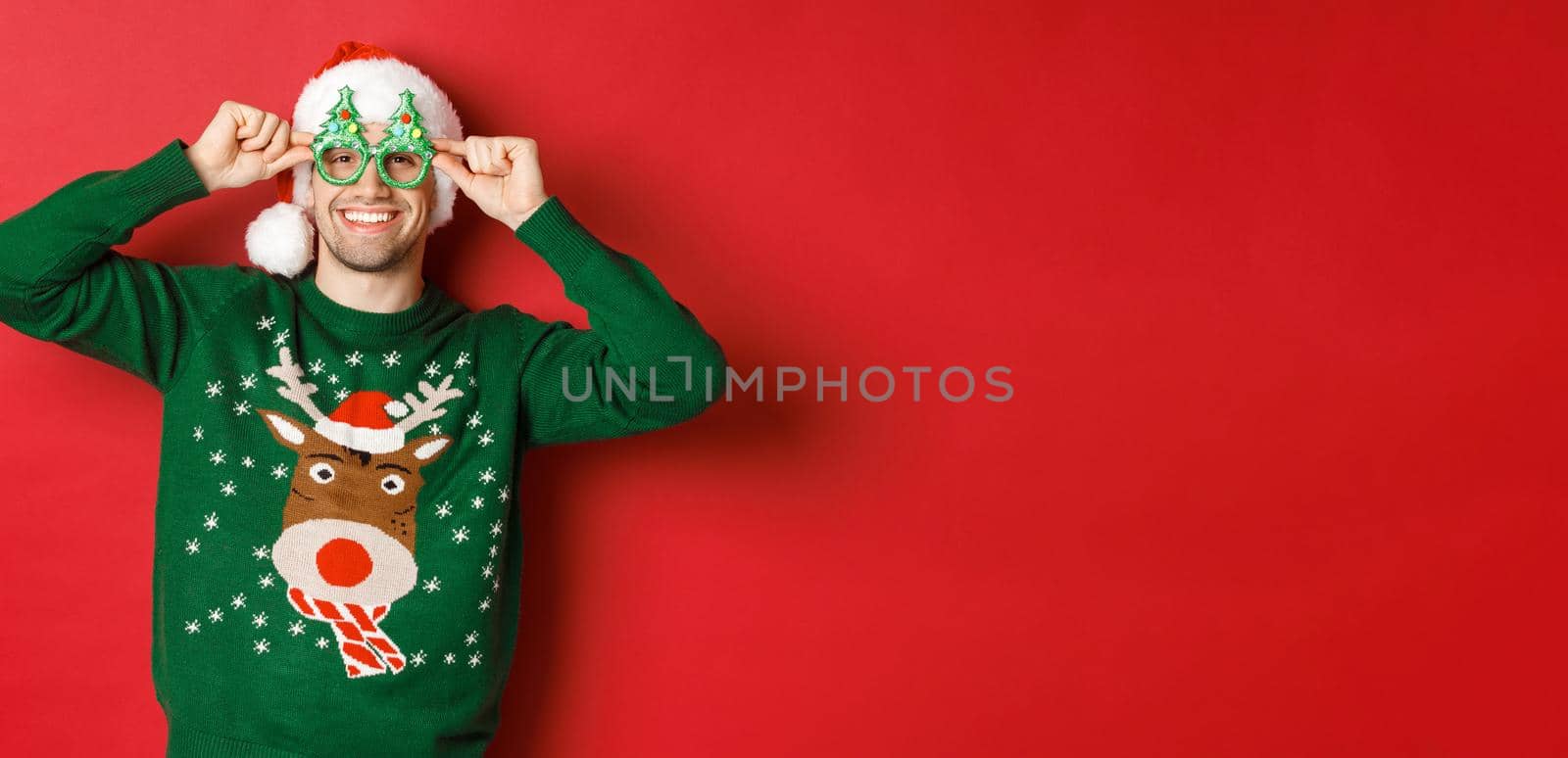 Portrait of attractive smiling man in santa hat, party glasses and sweater, celebrating new year holidays, standing against red background.
