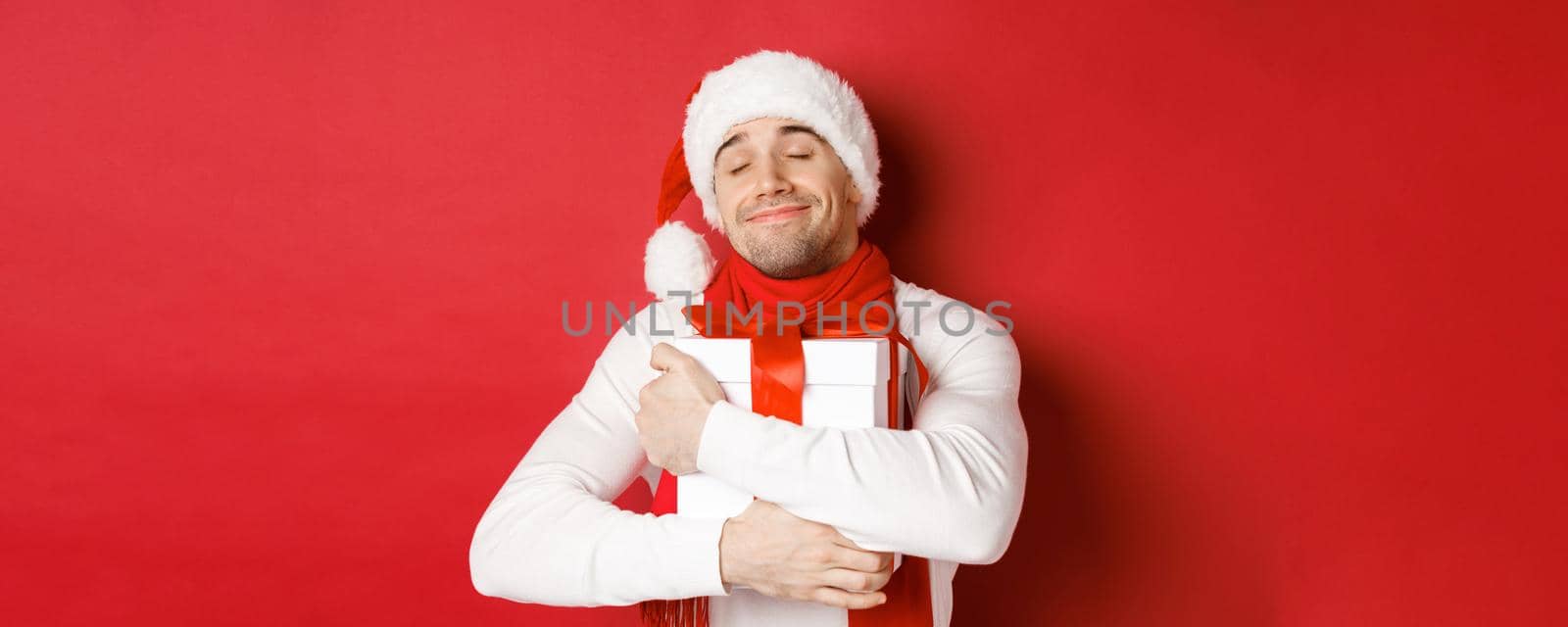 Concept of winter holidays, christmas and lifestyle. Image of lovely guy in santa hat and scarf, hugging his new year present and smiling flattered, standing over red background. Copy space