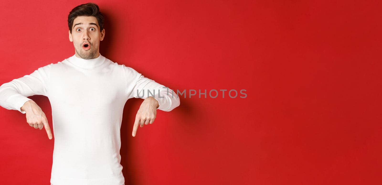 Image of surprised attractive guy in white sweater, showing announcement, pointing fingers down and looking amazed, standing over red background by Benzoix
