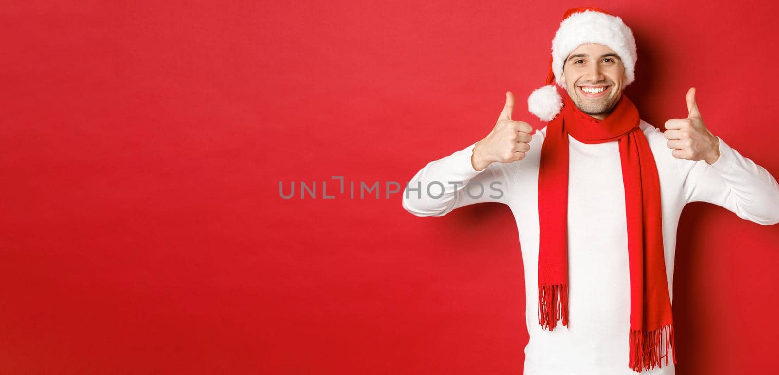 Portrait of handsome smiling man in santa scarf and hat, showing thumbs-up, celebrating christmas, standing over red background.