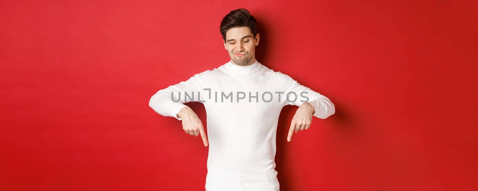Portrait of unamused handsome man, looking skeptical while pointing fingers down at something bad, standing over red background.