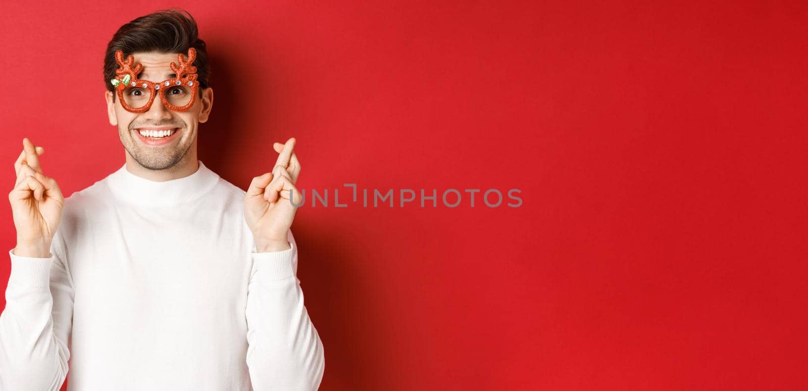 Close-up of excited handsome man, wearing party glasses and white sweater, crossing fingers for good luck, celebrating christmas and waiting wish, anticipating gifts, standing over red background.