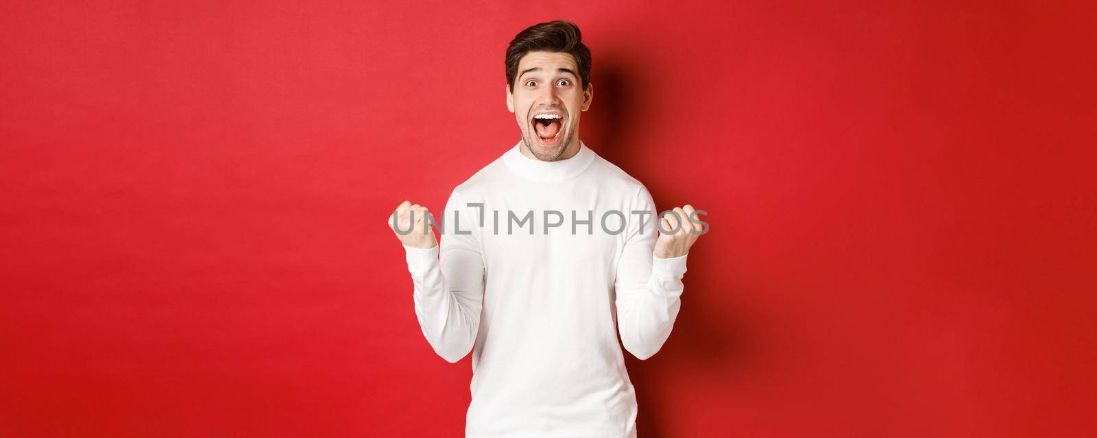 Image of happy good-looking man in white sweater, winning something, making fist pump and smiling amazed, celebrating victory, standing over red background by Benzoix