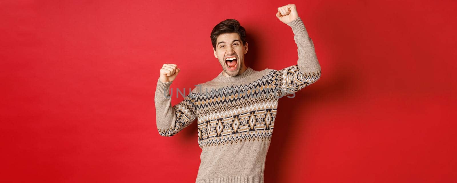 Image of happy handsome guy rejoicing on christmas eve, raising hands up and shouting for joy, triumphing and celebrating winter holidays, standing over red background by Benzoix
