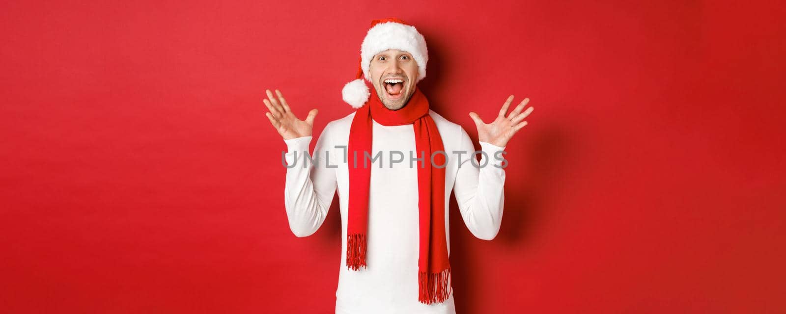 Portrait of happy and amazed handsome man, celebrating new year, wishing merry christmas, wearing santa hat and scarf, telling big news, standing against red background by Benzoix