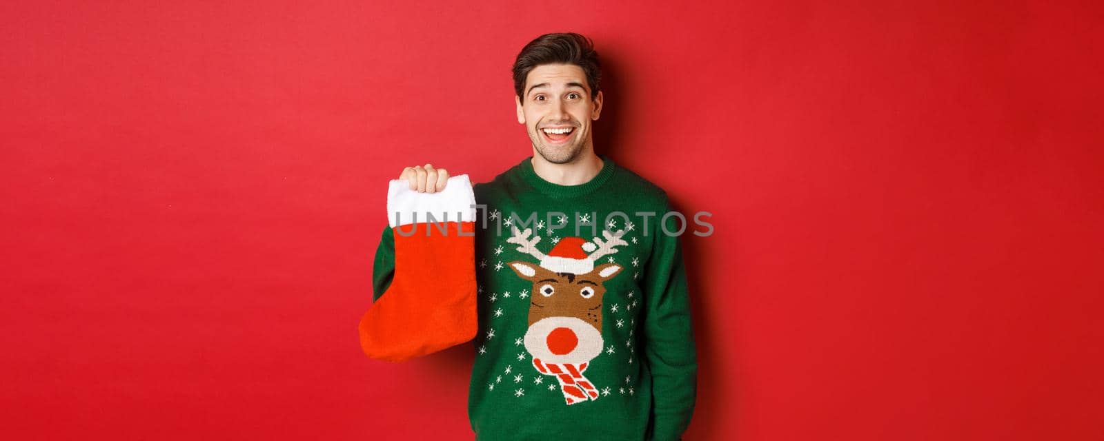 Image of handsome happy guy in sweater, holding christmas stocking and smiling amused, standing against red background by Benzoix