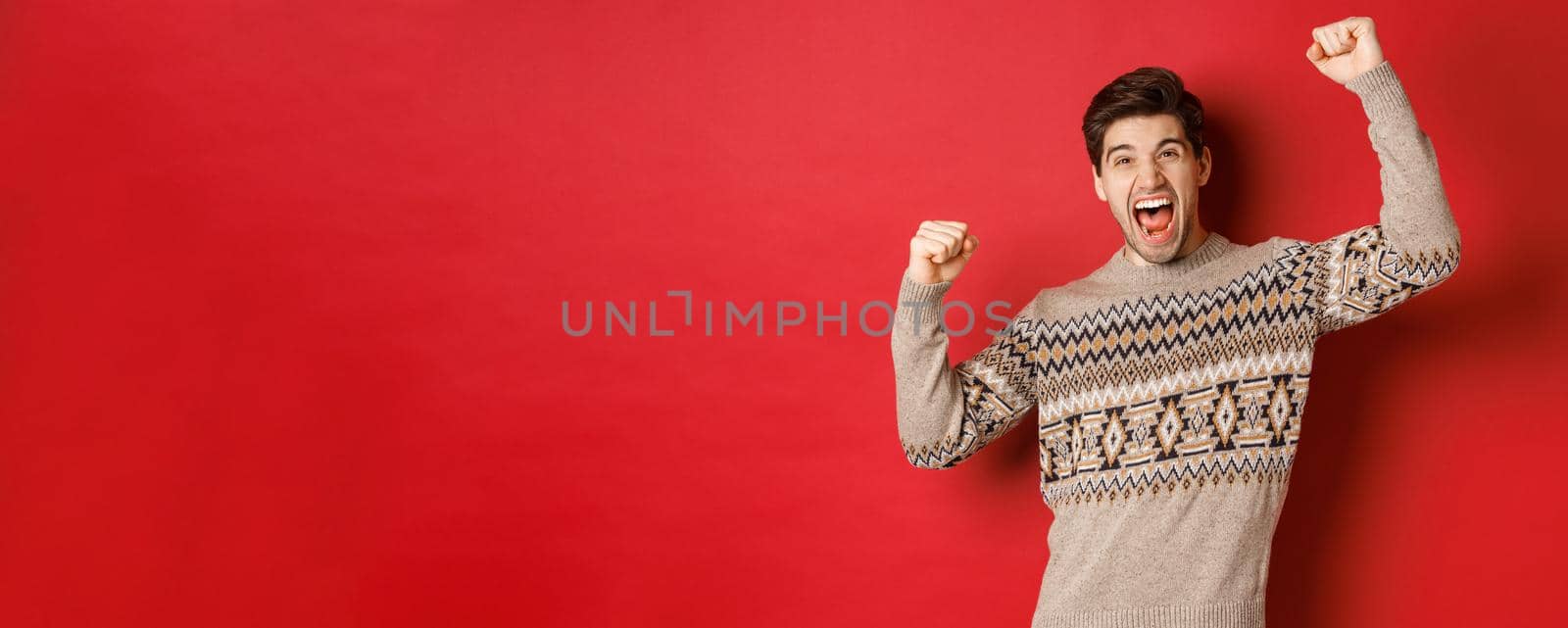 Image of happy handsome guy rejoicing on christmas eve, raising hands up and shouting for joy, triumphing and celebrating winter holidays, standing over red background.
