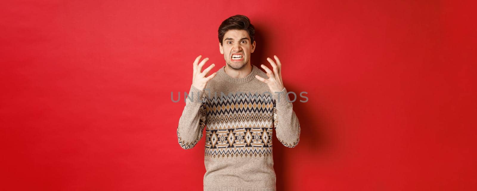 Image of angry and frustrated man in winter sweater, shouting and looking with hatred, standing upset over red background by Benzoix