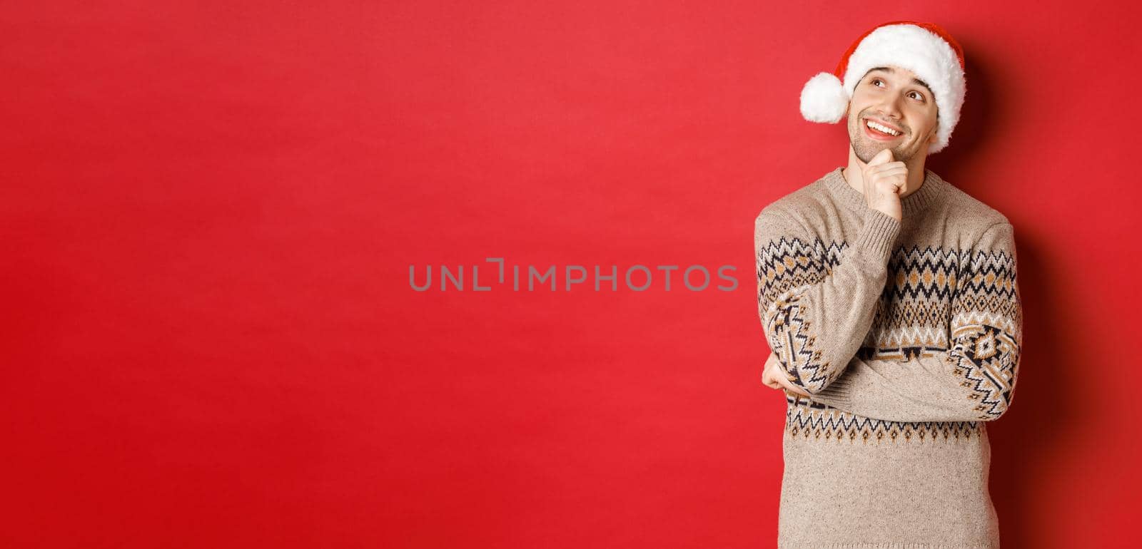 Image of happy young man in santa hat and christmas sweater, imaging something, thinking about new year gifts and smiling, looking at upper left corner, standing over red background by Benzoix