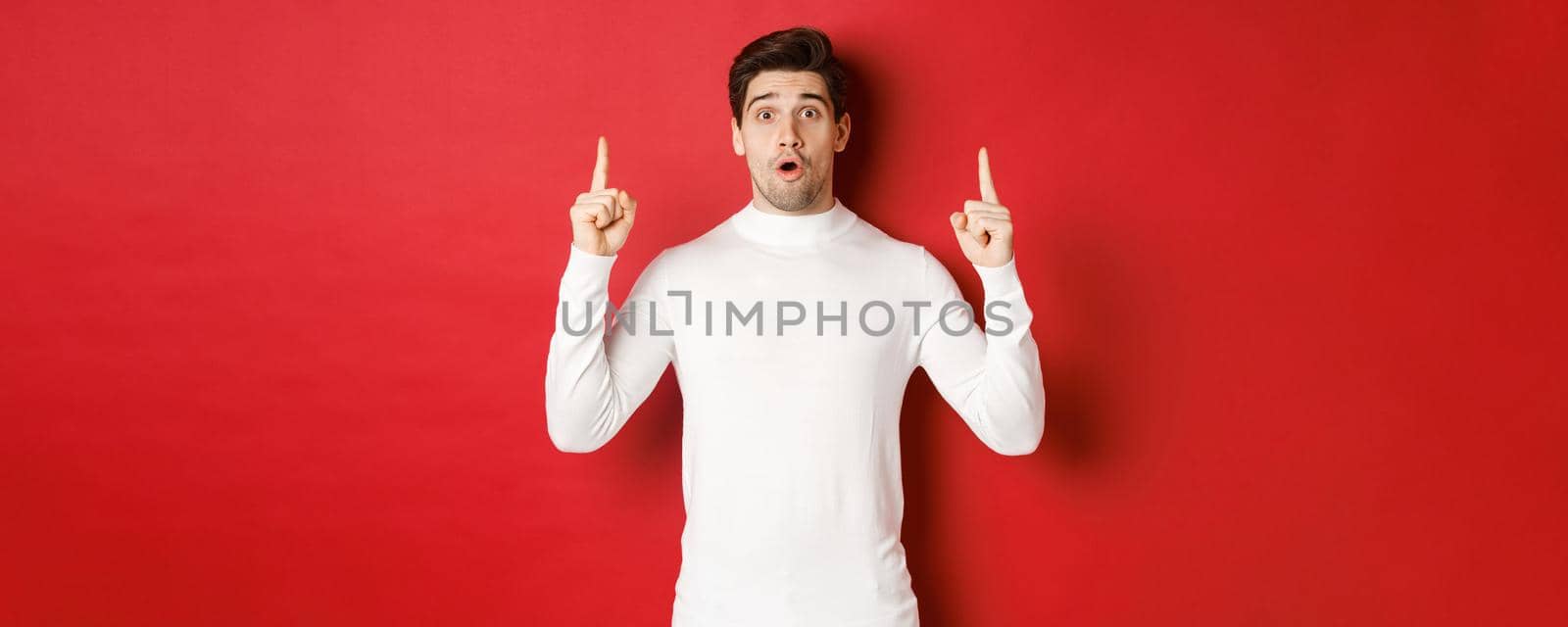 Concept of winter holidays. Good-looking young man with bristle, wearing white sweater, showing christmas advertisement on copy space, pointing fingers up and looking surprised, red background by Benzoix