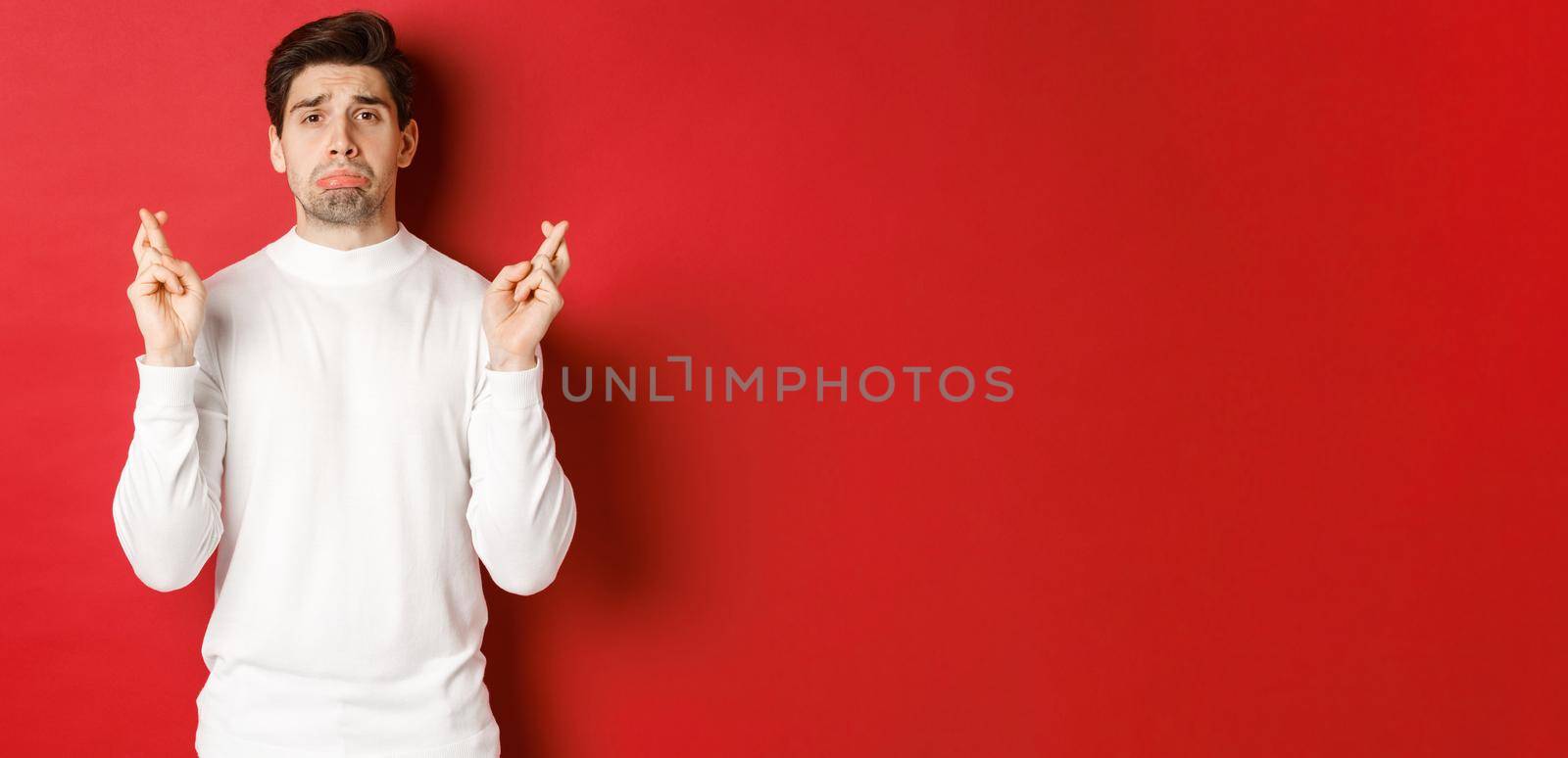 Image of sad and gloomy guy in white sweater, crying with fingers crossed, waiting for something or praying, standing over red background by Benzoix
