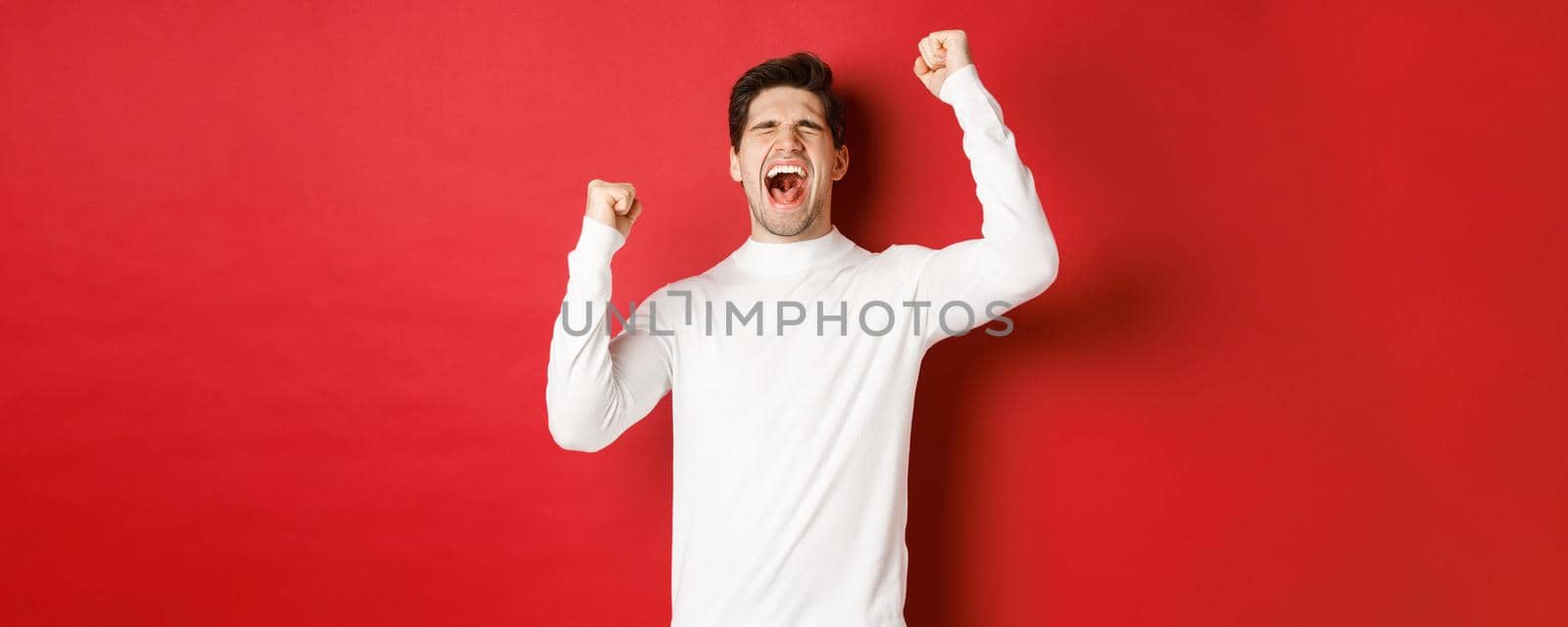 Portrait of handsome man in white sweater, feeling cheerful, celebrating victory, shouting for joy and raising hands up in victory, standing over red background by Benzoix