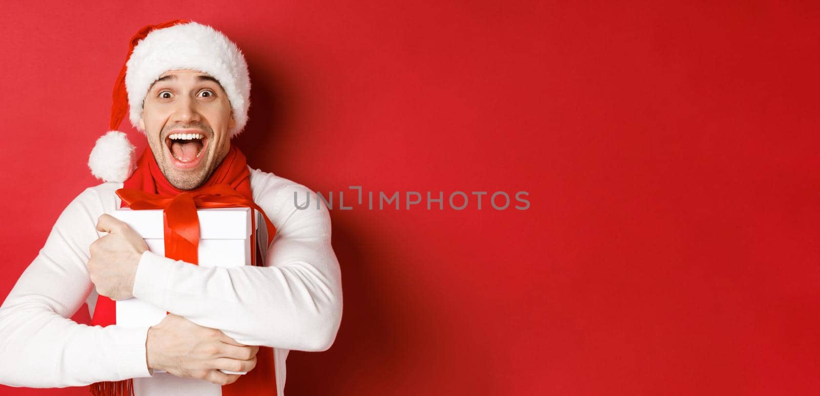 Concept of winter holidays, christmas and lifestyle. Close-up of happy man in santa hat receiving present, looking happy and hugging gift box, standing over red background by Benzoix